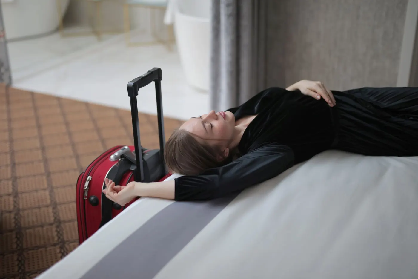 Una viaggiatrice esausta riposa sul letto di un albergo, accanto alla valigia